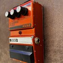 Kytarový efekt pedál v oranžové barvě s kovovým tělem. Na horní části jsou tři černé ovládací knoflíky a nápisy VOLUME, TONE, FUZZ. Uprostřed je bílý…