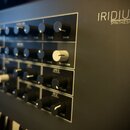 Detailní pohled na ovládací panel černého syntetizátoru s nápisem IRIDIUM SYNTHESIZER. Na panelu jsou vidět řady černých a bílých otočných potenciome…