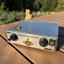 Stříbrný zesilovač v kovovém šasi s dvěma černými oválnými ovladači a zapínacím spínačem. Na předním panelu je nápis Little Mack Vintage Amp 1000, vl…