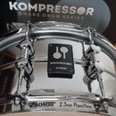 Stříbrný snare drum s chromovaným povrchem a obručemi. Na korpusu je černý čtvercový štitek s logem a nápisy KOMPRESSOR a SONOR. Na spodní obruči je…