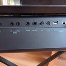 Detail zadní části černého elektronického nástroje s řadou konektorů. Jsou zde nápisy SUSTAIN, OUTPUT (s označením R a L/L+R), PHONES, MIC INPUT, GAI…