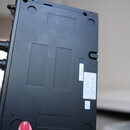 Fotografie zachycuje spodní stranu černého plastového elektronického zařízení. Na povrchu jsou vidět čtyři vlisované obdélníkové prohlubně a dvě kula…
