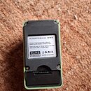 Zadní strana černého efektpedálu v zeleném rámu. Na zařízení je bílý štítek s nápisy AC ADAPTER DC9V, RoHS, CE a MADE IN CHINA, doplněný českým texte…
