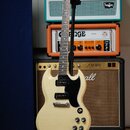 Elektrická kytara s bílým trupem a černým pickguardem umístěná na černém stojanu. Na hlavě hmatníku je viditelné logo Gibson. V pozadí jsou čtyři kom…