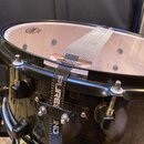 Černý buben s kovovými obručemi a napínacími šrouby. Přes membranu a korpus je přetažen šedý textilní popruh s nápisem SURESOUND. Na membraně je vidi…