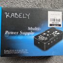 Fotografie znázorňuje krabici od napájecího zdroje s nápisem Multi-Power Supply. Na krabici je zobrazen černý zdrojem s nápisy POWER SUPPLY, IN DC 18…