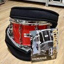Červený buben v černém polstrovaném obalu. Na koruse je vidět štítek se značkou YAMAHA a nápisem MODEL: MADE IN JAPAN. Před nástrojem leží produktový…