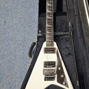 Bílá elektrická kytara s trojúhelníkovým tělem v černém tvrdém kufru. Na hlavě hmatníku je nápis Jackson. Nástroj má dva humbuckery, dva ovládací kno…