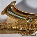 Detailní pohled na horní část zlatého saxofonu s lesklým povrchem. Na zvonu je vyryt nápis YAS-62 JAPAN a dekorativní ornamenty. Jsou vidět klapy s p…