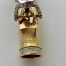 Detail zlatého kovového mouthpiece s nápisem DURGA na stříbrném kroužku u spodního konce. Nad nápisem je vyražen symbol osmičky v obdélníku a kód 7DG…