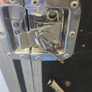 Detailní záběr na kovový zámek černého transportního kufru s hliníkovými hranami. Zámek je stříbrný, z kovu, s vloženým klíčem a vykazuje drobné škrá…