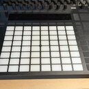 Černý MIDI kontrolér s mřížkou 8x8 bílých podsvícených padů. Zařízení obsahuje několik řad černých tlačítek, posuvníků a v horní části displej. Povrc…