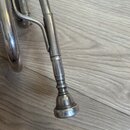 Detailní pohled na stříbrnou trubku z kovu. Je vidět část hmatníku a ozvukovka s viditelnými škrábanci a oxidací povrchu. Nástroj leží na světlém dře…