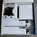 Fotografie ukazuje otevřenou bílou krabici s vložkami z polystyrenu a kartonu. Uvnitř je umístěn černý síťový adaptér s přiloženým kabelem a část dal…