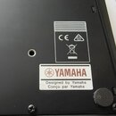 Detail spodní strany černého zařízení z plastu. Je zde vidět bílá nálepka s červeným logem a nápisem YAMAHA, pod kterým je text Designed by Yamaha a…