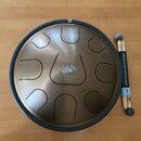 Kulatý steel tongue drum v bronzové barvě s černým okrajem. Uprostřed je logo a nápis ZENKO ionian. K nástroji jsou přiloženy dvě dřevěné paličky s g…