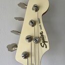 Záběr na hlavu basové kytary v krémové barvě. Na povrchu je černý nápis Squier by FENDER a JAZZ BASS®. Nástroj má čtyři chromované ladičky, z nichž j…