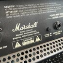 Detail zadní strany kytarového komba s černým povrchem. Je zde vidět bílý nápis Marshall a text Made in England by: Marshall Amplification plc, Bletc…