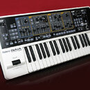 Bílo-černý syntetizátor s klávesnicí a ovládacím panelem s modrými, žlutými a oranžovými sekcemi. Na přední straně je nápis Roland GAIA SYNTHESIZER S…