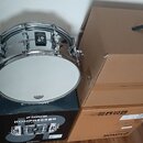 Stříbrný snare drum s bílým koženem, umístěný na černé krabici. Na obruči je černý oválný prvek s bílým symbolem a na koženě logo SONOR. Černá krabic…
