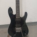 Černá elektrická kytara s tmavým hnedým hmatníkem a dvěma humbuckery. Nástroj je umístěn ve stojanu a má připojený stříbrný tremolo páku. Na těle nej…