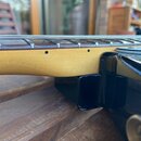 Detailní pohled na spoj hned krku a těla elektrické kytary. Krk je vyroben ze světlého dřeva s viditelnou strukturou, tělo je černé s vysokým leskem.…