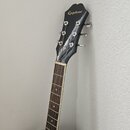Černá akustická kytara s hnedým hmatníkem. Na hlavě je zlatý nápis Epiphone a šest chromovaných ladiček. Povrch nástroje je hladký a v dobrém stavu.