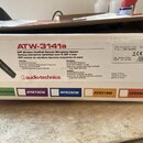 Fotografie znázorňuje kartónovou krabici od bezdrátového mikrofonního systému. Na bílém štítku je uveden model ATW-3141a a značka audio-technica. Tex…