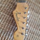 Fotografie zachycuje hlavu elektrické kytary z přírodního dřeva. Na povrchu je černé logo Fender a nápisy STRATOCASTER, MADE IN JAPAN s textem WITH S…