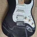 Elektrická kytara typu Stratocaster s černým průsvitným lakem, pod kterým je vidět struktura dřeva. Nástroj má bílý perleťový pickguard, javorový hma…