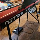 Červené digitální piano na černém stojanu s připozeným pedálem. Na boku nástroje je bílý nápis nord piano. Povrch červeného šasi vykazuje drobné oděr…