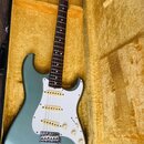 Modrošedá elektrická kytara typu Stratocaster s bílým pickguardem a hnědým hmatníkem v žlutém plišovém kufru. Na hlavě je vidět logo Fender. Nástroj…
