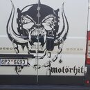 Na fotografii je zadní část bílého vozu Peugeot Boxer. Na dveřích je velká černá grafika s vyobrazením lebky v přilbici a nápis motörhit. Vozidlo má…