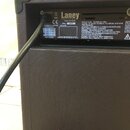 Detail zadní strany hnědého kytarového komba s texturovaným povrchem. Na černém štítku je bílý nápis Laney, pod ním text Power To The Music a adresa…