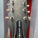 Detail hlavy elektrické kytary v černé barvě. Na horní části je zlatý nápis Gibson a uprostřed svisle orientovaný nápis Les Paul MODEL. Dole se nachá…