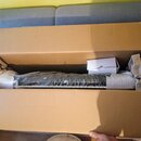 V otevřené kartonové krabici s polystyrenovými výplněmi leží hudební nástroj zabalený v průhledné plastové fólii. Vedle nástroje je umístěna menší bí…