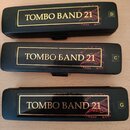 Tři černé harmoniky s nápisem TOMBO BAND 21 v zlatém rámečku. Každý nástroj má v pravém horním rohu označení tóniny: D, C a G. Na tělech jsou vidět n…