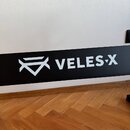 Detailní záběr na černý povrch s bílým nápisem VELES-X a doprovodným geometrickým logem. Povrch je hladký, matný a v dobrém stavu. Pod černým prvkem…