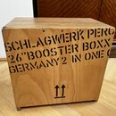 Dřevěný nástroj ve tvaru kostky s přirozenou barvou dřeva. Na přední straně je černý nápis SCHLAGWERK PERC 26"BOOSTER BOXX GERMANY2 IN ONE O a ve spo…