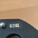 Detailní záběr na černý povrch hudebního vybavení. Na povrchu je nalepený černý štítek s bílým nápisem IS-1216SL. Vedle štítku je vidět kovový šroub…