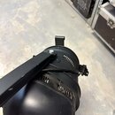 Černý bodový reflektor z kovového materiálu s připevněným držákem a úchytem. Povrch je matný s viditelnými prachovými částicemi a drobným opotřebením…
