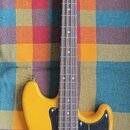 Žlutá elektrická basová kytara s černým pickguardem a hnedým hmatníkem. Na hlavě je svisle napsaný nápis "Precision Bass". Nástroj vykazuje známky po…