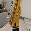 Fotografie zachycuje hlavu elektrické kytary v žluté barvě s hnedým hmatníkem. Na povrchu je černý nápis Fender® a svislý text TELECASTER®. Jsou vidě…