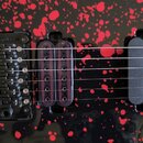 Detail elektrické kytary s černým povrchem a červenými stříkanci. Je zde vidět černý kobylník s nápisem FloydRose, humbucker s fialovými cívkami a da…