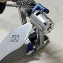 Detailní pohled na kovový mechanismus bubnového pedálu v stříbrné a modré barvě. Na stříbrném šlapadle je vyryto logo tvořené dvěma protilehlými troj…