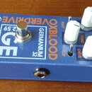 Modrý efekt pedál s bílým a oranžovým nápisem. Na horní straně jsou vidět texty OXBLOOD, GERMANIUM 32, VERDRIVE a GE, dále nápisy PHAT, TONE, CLIP a…