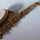 Zlatý saxofon s kovovým povrchem, položený na bílém textilu. Nástroj je kompletní s nasazeným moutkem, bez viditelných značek, log nebo modelových oz…