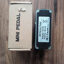 Na fotografii je malý efektový pedál v modré barvě a jeho hnědá kartonová krabice s nápisem MINI PEDAL. Na spodní straně pedálu je bílý štítek s text…