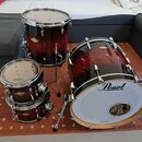 Sada bubnů v tmavě červeném průsvitném provedení s viditnou strukturou dřeva. Na basovém bubnu je bílý přední kožen s nápisem Pearl a malým logem Mas…