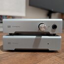 Dvě stříbrné kovové krabičky v tzv. stacku umístěné na dřevěném povrchu. Horní zařízení má nápis MAGNI s logem H, ovládací knoflík a konektor pro slu…
